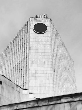 A large, modernist building with a distinctive pyramid-like shape, featuring a circular black emblem near the top. The structure is covered with numerous rectangular tiles, creating a textured facade. The architecture gives a sense of monumentalism and simplicity.