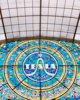 A colorful stained glass window featuring the word 'TESLA' prominently in the center, surrounded by a circular pattern with geometric shapes in shades of blue, yellow, and red. The design is intricate and symmetrical, showcasing detailed craftsmanship.