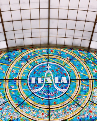 A colorful stained glass window featuring the word 'TESLA' prominently in the center, surrounded by a circular pattern with geometric shapes in shades of blue, yellow, and red. The design is intricate and symmetrical, showcasing detailed craftsmanship.