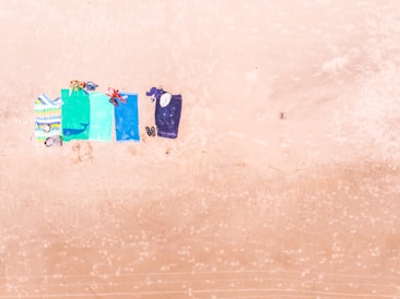 Five beach towels are spread out on the sand next to each other. Each towel is a different color or pattern, and various personal items such as shoes, hats, and swimwear are placed on or near the towels.