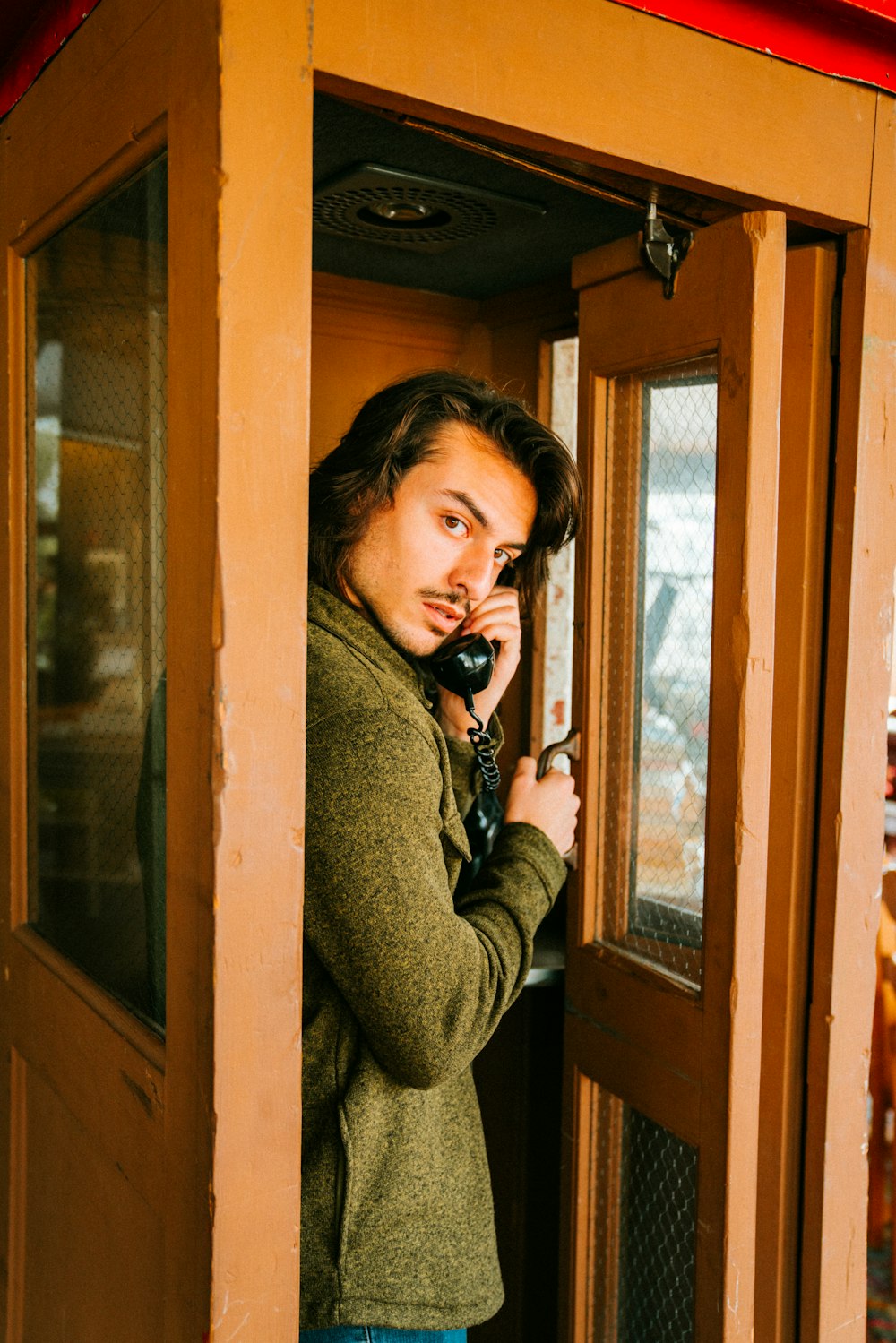 man in brown jacket standing inside phone boot and holding phone