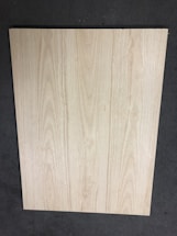 A large, rectangular piece of light wood with prominent grain patterns rests on a dark, textured floor. The wood surface appears smooth and flat with natural coloration and subtle variations in the wood grain.
