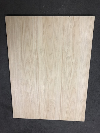 A large, rectangular piece of light wood with prominent grain patterns rests on a dark, textured floor. The wood surface appears smooth and flat with natural coloration and subtle variations in the wood grain.