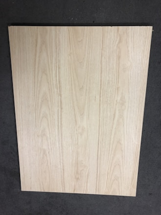 A large, rectangular piece of light wood with prominent grain patterns rests on a dark, textured floor. The wood surface appears smooth and flat with natural coloration and subtle variations in the wood grain.