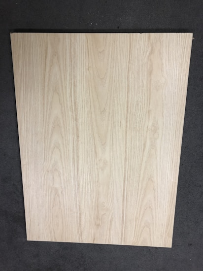 A large, rectangular piece of light wood with prominent grain patterns rests on a dark, textured floor. The wood surface appears smooth and flat with natural coloration and subtle variations in the wood grain.