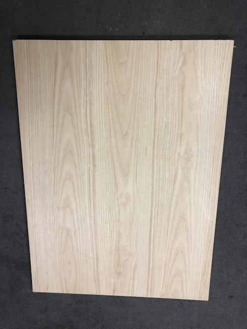 A large, rectangular piece of light wood with prominent grain patterns rests on a dark, textured floor. The wood surface appears smooth and flat with natural coloration and subtle variations in the wood grain.