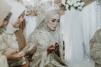A group of women dressed in intricately embroidered hijabs and traditional attire are sitting in a solemn and focused posture. The main woman in the center is wearing a detailed gown with a sparkling tiara. They appear to be engaged in a prayer or blessing ceremony, with their hands raised in a cupped gesture. A backdrop of white flowers and soft-colored drapery creates an elegant and serene setting.
