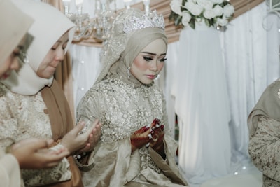 A group of women dressed in intricately embroidered hijabs and traditional attire are sitting in a solemn and focused posture. The main woman in the center is wearing a detailed gown with a sparkling tiara. They appear to be engaged in a prayer or blessing ceremony, with their hands raised in a cupped gesture. A backdrop of white flowers and soft-colored drapery creates an elegant and serene setting.