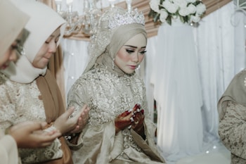A group of women dressed in intricately embroidered hijabs and traditional attire are sitting in a solemn and focused posture. The main woman in the center is wearing a detailed gown with a sparkling tiara. They appear to be engaged in a prayer or blessing ceremony, with their hands raised in a cupped gesture. A backdrop of white flowers and soft-colored drapery creates an elegant and serene setting.