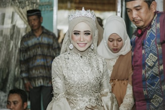 A bride in an ornate light-colored dress with intricate floral detailing and a sparkling tiara is smiling. She is surrounded by people dressed in traditional garments, capturing a sense of cultural celebration.