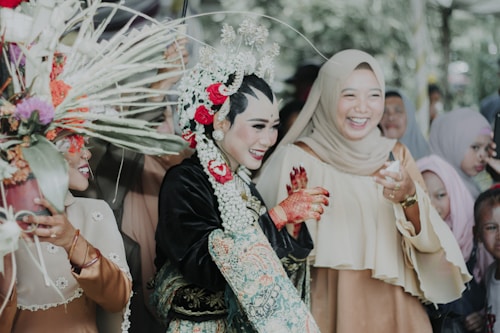 A joyful group of people is gathered at a cultural event. A person dressed in elaborate traditional attire, adorned with floral and intricate headpieces, is at the center. The surrounding individuals are smiling and appear to be celebrating. The atmosphere is festive, with colorful decorations.