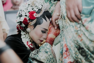 A woman dressed in traditional attire with intricate floral hair decorations and detailed jewelry bows her head in a gesture of respect or reverence. Her hands, adorned with red henna patterns, hold onto a fabric, possibly part of another person's clothing, who stands nearby.