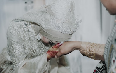 A person wearing an intricately detailed, embellished garment and headpiece is bending down to kiss another person's hand, which is adorned with red henna patterns. The second person is wearing a light-colored embroidered outfit.