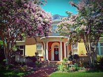 A beautiful two-story brick house with classic architectural elements, surrounded by lush greenery and large, blooming crepe myrtle trees with pink flowers. The central entrance features a dark wooden door with a circular design, flanked by two white columns that add to its elegant facade. The windows are detailed with diamond lattice patterns, and the garden is well-maintained with various shrubs and plants.