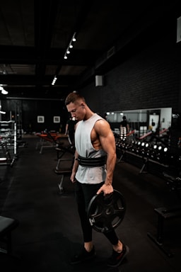 A muscular person is seen standing in a gym, holding a large weight plate in one hand. They are wearing a sleeveless white shirt and black workout pants, with a weightlifting belt around their waist. The gym is dimly lit, with equipment and mirrors visible in the background.