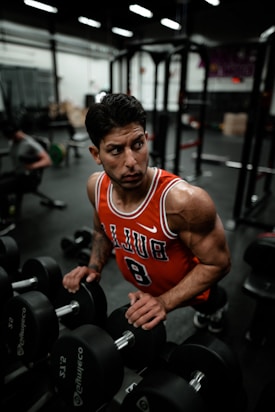 A muscular man in an orange sleeveless jersey with the number 8 is standing in a gym, gripping a dumbbell rack. He has a focused and intense expression as he looks to his left. The background shows gym equipment and another individual working out.