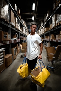 A person stands in a warehouse aisle holding large yellow shopping bags. The setting is industrial with shelves lined with boxes and items. Overhead lighting creates shadows, adding a dramatic effect.