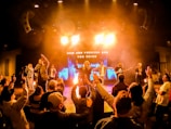 A vibrant concert scene with a band performing on stage under bright lighting. The audience is engaged, raising their hands, and appears to be enjoying the music. The background features an illuminated display with the text 'Now and Forever God You Reign'.