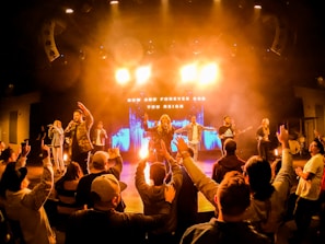 A vibrant concert scene with a band performing on stage under bright lighting. The audience is engaged, raising their hands, and appears to be enjoying the music. The background features an illuminated display with the text 'Now and Forever God You Reign'.