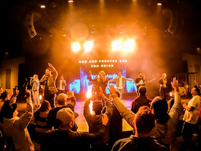 A vibrant concert scene with a band performing on stage under bright lighting. The audience is engaged, raising their hands, and appears to be enjoying the music. The background features an illuminated display with the text 'Now and Forever God You Reign'.