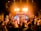 A vibrant concert scene with a band performing on stage under bright lighting. The audience is engaged, raising their hands, and appears to be enjoying the music. The background features an illuminated display with the text 'Now and Forever God You Reign'.