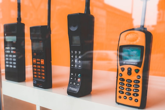 A collection of four vintage mobile phones displayed in a row against an orange background. The phones vary in size and shape, showcasing different designs from past eras, including prominent antennas and tactile keypads.