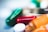 An artistic close-up of various colorful pills, featuring a red capsule, a teal gel pill, and a white round tablet. The image is softly focused, giving a dreamy or abstract feel with radiant highlights and bokeh in the background.