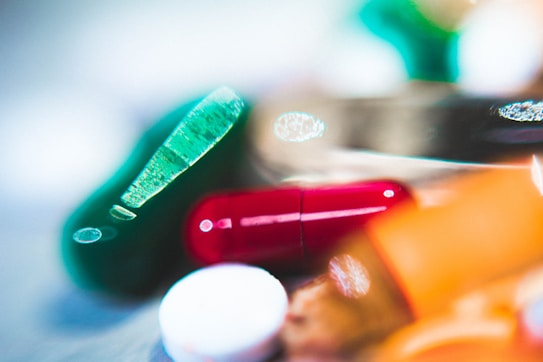 An artistic close-up of various colorful pills, featuring a red capsule, a teal gel pill, and a white round tablet. The image is softly focused, giving a dreamy or abstract feel with radiant highlights and bokeh in the background.