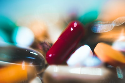 A close-up view of various colorful pills and capsules scattered across a surface. The image is out of focus with a dreamy, abstract quality, emphasizing the vibrant reds, greens, oranges, and whites of the medication.