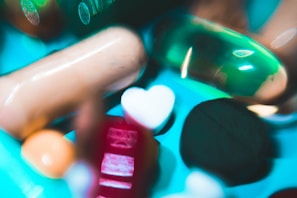 A collection of various pills and capsules, including a prominent green gel capsule and a heart-shaped white tablet. The image has a colorful and slightly abstract presentation with a blurred, artistic style.
