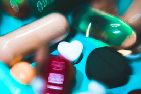 A collection of various pills and capsules, including a prominent green gel capsule and a heart-shaped white tablet. The image has a colorful and slightly abstract presentation with a blurred, artistic style.