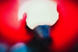 A blurred image with a dominant bright red background, a central white circular light, and hints of dark blue and black shapes that are not clearly identifiable.