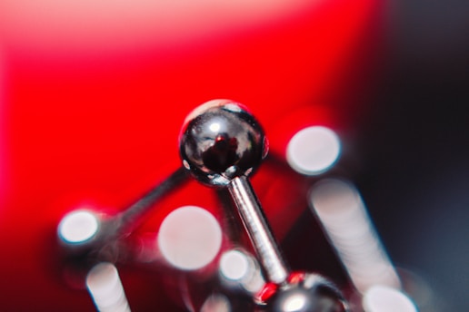 A close-up of a shiny, metallic structure that resembles a molecular model. The background features a vibrant red hue, with bokeh circles of light scattered across the image, creating an abstract, artistic feel.