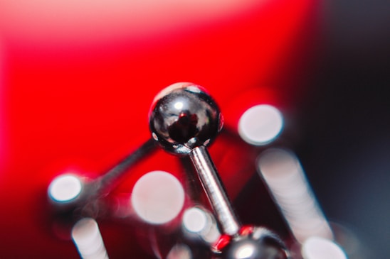 A close-up of a shiny, metallic structure that resembles a molecular model. The background features a vibrant red hue, with bokeh circles of light scattered across the image, creating an abstract, artistic feel.