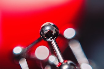 A close-up of a shiny, metallic structure that resembles a molecular model. The background features a vibrant red hue, with bokeh circles of light scattered across the image, creating an abstract, artistic feel.