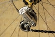 A close-up of a bicycle gear mechanism, showcasing a Shimano derailleur attached to the rear wheel. The image highlights the metallic components and tensioned chain against a neutral background. The bicycle spokes and other gear parts are also visible.
