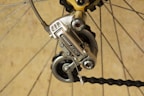 A close-up of a bicycle gear mechanism, showcasing a Shimano derailleur attached to the rear wheel. The image highlights the metallic components and tensioned chain against a neutral background. The bicycle spokes and other gear parts are also visible.