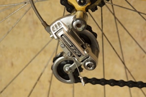 A close-up of a bicycle gear mechanism, showcasing a Shimano derailleur attached to the rear wheel. The image highlights the metallic components and tensioned chain against a neutral background. The bicycle spokes and other gear parts are also visible.