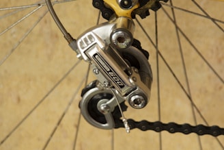 A close-up of a bicycle gear mechanism, showcasing a Shimano derailleur attached to the rear wheel. The image highlights the metallic components and tensioned chain against a neutral background. The bicycle spokes and other gear parts are also visible.
