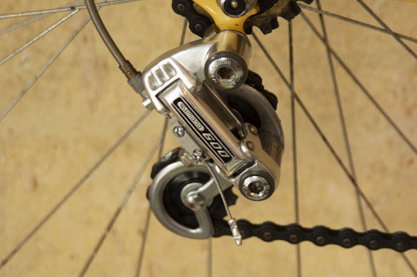A close-up of a bicycle gear mechanism, showcasing a Shimano derailleur attached to the rear wheel. The image highlights the metallic components and tensioned chain against a neutral background. The bicycle spokes and other gear parts are also visible.