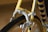 A close-up view of a bicycle brake system on a yellow frame, highlighting the metallic brake caliper and cable. The tire and part of the rim are visible, with spokes in the background. The frame features a decorative pattern of stripes in multiple colors.