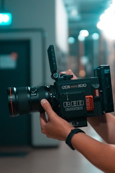 A professional camera is being held by a person wearing a black wristband. The camera is marked with the words 'RED RAVEN' and has a large lens attached. The background is blurred, with soft lighting and a mix of dark and light areas, suggesting an indoor setting.