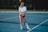 A person is standing on a tennis court with a net in the background, wearing an outfit consisting of a white crop top, red shorts, high socks, and sneakers. The individual is holding a tennis racket and appears relaxed.