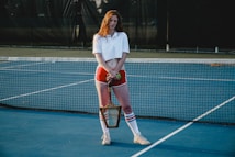 A person is standing on a tennis court with a net in the background, wearing an outfit consisting of a white crop top, red shorts, high socks, and sneakers. The individual is holding a tennis racket and appears relaxed.
