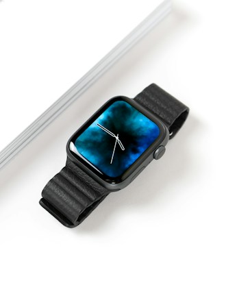 A sleek smartwatch with a black leather strap and a vivid blue, cloud-like design on its rectangular display. The face shows a classic analog clock design with minimalist white hands. It rests on a white surface, next to a hardcover book with visible pages.
