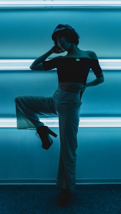 A person strikes a pose in front of bright, horizontal neon lights. The lighting creates strong silhouettes and contrasts, emphasizing the stylish, fashionable attire including a cropped top, wide-legged pants, and platform shoes. The scene has a modern and edgy feel.