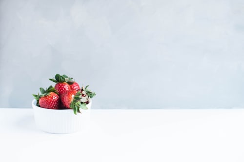 Four red strawberries in white bowl, sitting on a white table. Background is light blue. Tacoma weed. Tacoma marijuana. Tacoma dispensary.