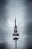 A tall telecommunication tower with various circular platforms below the antenna stands prominently against a backdrop of dramatic, overcast clouds. The tower's structure features a mix of white and dark sections, with red accents on the antenna.
