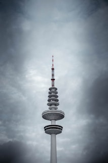 A tall telecommunication tower with various circular platforms below the antenna stands prominently against a backdrop of dramatic, overcast clouds. The tower's structure features a mix of white and dark sections, with red accents on the antenna.