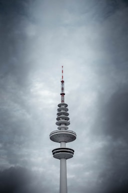 A tall telecommunication tower with various circular platforms below the antenna stands prominently against a backdrop of dramatic, overcast clouds. The tower's structure features a mix of white and dark sections, with red accents on the antenna.